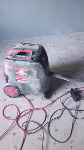 A person in a red t-shirt and yellow shorts is vacuuming a room with a long hose attached to a vacuum cleaner. The room has tiled floors and gray walls. There is a window with blinds on the right side of the room. The ceiling is made of wood panels. A black vacuum cleaner is visible in the foreground, connected to the hose by a power cord. The activity appears to be a cleaning or renovation project. The time of day is not specified, and there are no clear weather cues. The location is identified as Ladyzhyn, Ukraine. No visible text is present in the media file.