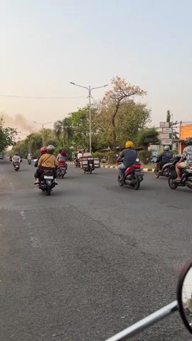 A group of motorcyclists are riding down a paved road. The motorcyclists are of various ages and genders, and they are riding a variety of motorcycles. Some of the motorcycles are carrying passengers. The road is lined with trees and bushes. There is a lamppost in the background. The sky is hazy and overcast.