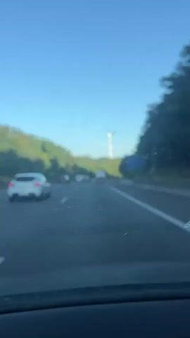 The video captures a view from inside a car driving on a highway. The road ahead is relatively clear, with several cars visible at a distance. On the left side of the highway, there are green, tree-covered hills. On the right side, there is a sign. Further in the distance, a large white wind turbine is visible against a clear blue sky. The sun appears to be shining, casting a warm light on the scene. The perspective suggests the car is moving forward at a moderate pace.