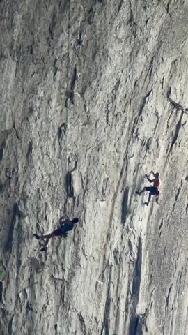 The video shows two people rock climbing up a large, gray rock face, with ropes and harnesses. The camera zooms out to reveal that they are climbing under a massive natural archway, with a bright blue sky visible through the opening. Trees and vegetation grow at the base of the archway. The perspective is from below, looking up at the climbers.