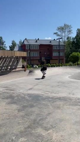 The video shows a person on a skateboard performing a trick in a skatepark. The person is wearing a black t-shirt and white shorts and is captured mid-air in an upside-down position, with their skateboard beneath them. The skatepark appears to be made of concrete and features ramps and bowls. In the background, there is a multi-story brick building with large windows and a blue sky overhead. Trees are visible behind the building. The lighting suggests it is daytime.