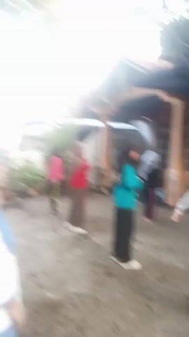 The video shows a group of people standing in what appears to be an outdoor area, possibly a schoolyard or a residential compound. The image quality is poor, with significant motion blur, making it difficult to discern fine details.

In the first clip, several individuals are visible, some wearing bright clothing like red and pink. They appear to be standing in a line or a group. The background shows what could be houses with tiled roofs and trees.

The second clip shifts to a different scene, again featuring several people from behind. One person in the foreground is wearing a yellow top, and another in the middle ground is in a pink top and dark pants. They are standing in front of a building with a large, dark entrance.

The third clip shows a person wearing a light blue hijab and a dark skirt. They are standing in an outdoor area with visible debris on the ground. Sunlight appears to be casting a strong glare, further obscuring details. Other individuals are also present in the background, though their features are indistinguishable due to the blur and lighting. The overall impression is of an informal gathering or activity taking place outdoors.