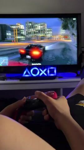 The video shows a person playing a driving game on a TV. The game, which appears to be Grand Theft Auto, is displayed on a large flatscreen TV. The player is using a PlayStation controller, which is red and black. The TV sits on a white table, and a blue PlayStation symbol light is positioned in front of the screen.  The person's legs are visible, clad in black shorts with a cartoon character on them. The room is dim, illuminated by the TV and the PlayStation light.