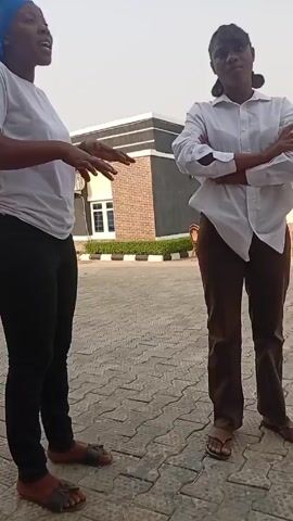 Two women stand in front of a building on a paved lot. One woman wears a white t-shirt, black pants, and brown sandals. She also wears a blue head wrap. The other woman wears a white button-down shirt, brown pants, and brown sandals. She has her arms crossed. The two women appear to be having a discussion. The camera angle is from low to high and remains stationary.