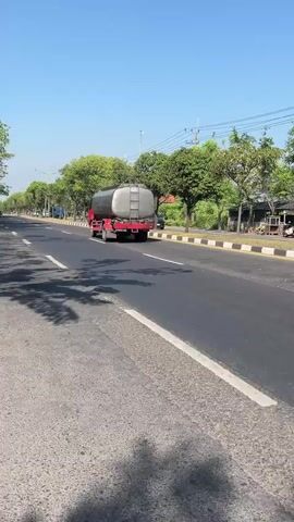 The media file captures a wide shot of a road in Ketapang, Indonesia, on a clear, sunny day. The main subject is a red tanker truck with a silver cylindrical tank, traveling away from the camera on the right side of the asphalt road. The truck appears to be a commercial vehicle, likely transporting liquids.

The setting is a relatively straight road lined with trees on both sides. On the right side of the road, there's a sidewalk with a yellow and black striped curb, followed by some low buildings and more trees. On the left side, the road continues with a white lane marking and a shadow of trees stretching across the asphalt, indicating the sun is to the left and slightly behind the viewer. In the distance, more vehicles and trees are visible, suggesting a somewhat rural or suburban environment. The sky is a bright, clear blue, indicative of good weather.

No people are clearly discernible in the frame. The activity is simply traffic flow, with the tanker truck being the most prominent vehicle in motion. Notable details include the bright sunlight casting distinct shadows, the uniform blue sky, and the green foliage of the trees. The overall mood is peaceful and ordinary, depicting a typical scene on a roadway. There is no visible text within the frame.