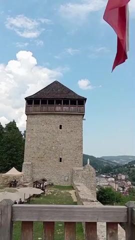 The video begins with a view of a stone tower with a wooden roof against a cloudy blue sky. A red and white flag waves in the foreground. The camera pans right, revealing a town nestled in a valley with hills and forests in the background. A river flows through the town, spanned by a bridge. The camera continues to pan, showing more of the town and surrounding landscape, including roads, buildings, and trees. The video ends with a panoramic view of the town and its natural surroundings.