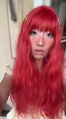 A person with pale-toned skin, red wig with bangs, and pink and white makeup around the eyes and cheeks, poses for a selfie. The person is wearing a white top with ruffles. The hair has some star and lightning bolt clips. A window is visible in the background.