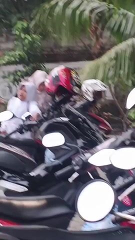 The video shows a parking area in front of a building with text on it that reads "SHOLIA AT-TA WA". Several motorcycles are parked, and a few people are visible. In the foreground, two motorcycles are partially obscured. Further back, a silver car is parked. A young child is sitting in the car. Another child is visible near the car. Two men are sitting under an overhang in front of the building, and one woman is seen walking. One of the men is wearing a blue shirt and the other a grey shirt. Another person, possibly a woman wearing a hijab, is seated near the entrance of the building. The environment appears to be outdoors with some trees and foliage in the background.