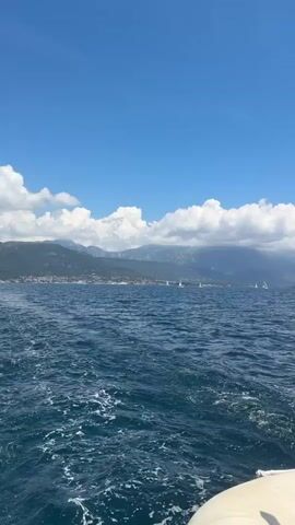 The video captures a scenic view from a moving boat on deep blue water, showing a white wake trailing behind. A majestic range of mountains stretches across the distant horizon, their peaks partially obscured by large, fluffy white and grey clouds against a bright blue sky. Several white sailboats are visible on the water, appearing to be actively sailing. As the video progresses, the camera zooms in slightly, bringing the sailboats and the cloud-covered mountains into sharper focus. A small red buoy is also visible in the mid-ground. The scene depicts a bright, sunny daytime environment off the coast of Bijela, Montenegro, with clear visibility and pleasant weather conditions for sailing.