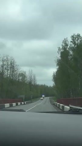A car is driving over a bridge on a rural road on a cloudy day. The road is flanked by trees, and in the distance, we can see an ambulance approaching.