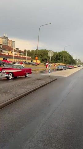 A slow-moving procession of vintage cars, including a red convertible with white-wall tires, proceeds along a wet asphalt road. The cars are lined up on the right side of the road, indicating a parade or gathering. On the left side of the road, a building with yellow and white siding and several people are visible. Trees form a backdrop behind the building and stretch along the right side of the road in the distance. The sky is overcast and grey. The camera appears to be mounted on a moving vehicle, capturing the scene from a low angle.