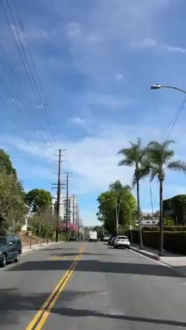 The video shows a street scene on a sunny day with a blue sky dotted with clouds. The street has two yellow lane markings and power lines overhead.  Along the sides of the street are parked cars, trees, and buildings. A white vehicle travels in the distance. The street is labeled "SLOW SCHOOL" in yellow paint.