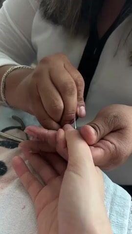 In a close-up indoor shot, two people are engaged in a manicure session. One individual, with darker skin tone, wearing a white shirt with a black strap and a silver bracelet on their left wrist, is carefully holding the hand of another person with lighter skin tone. The manicurist is using a pair of metal cuticle nippers to work on the fingernail and cuticle area of the other person's index finger. A white towel and a light blue towel are visible beneath the hands, indicating a prepared workspace. The focus is entirely on the hands and the intricate nail care process. No faces or specific location cues are visible.