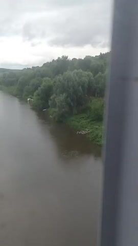 The video shows a view of a village from a moving vehicle, possibly a train. The village is nestled amongst rolling green hills covered in trees. A river runs alongside the village, and there are several houses visible, most of them appearing to be made of wood with red or brown roofs. Some of the houses have small yards with greenery. The sky is overcast and the overall atmosphere is calm and rural. The video is shot from a slightly elevated perspective, giving a panoramic view of the landscape.