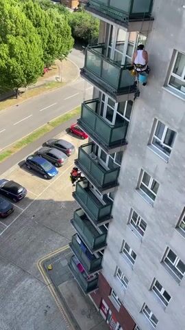 The video shows two men working on the exterior of a tall apartment building. They are suspended by ropes, presumably cleaning or repairing the building's facade. The building is multiple stories high and has green-railed balconies on each floor. The balconies are stacked vertically, creating a distinctive pattern on the building's exterior.  A parking lot is visible below with several cars, including a red one, parked in it. A street runs alongside the building with trees lining the opposite side. The sky is bright and the overall scene appears to be well-lit. One worker is higher up, wearing a white tank top and helmet, and has a bucket attached to his gear. The other worker is lower down and is wearing an orange jumpsuit and helmet.