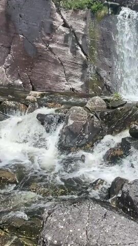 This is a vertical, medium shot of a cascading waterfall flowing over dark, wet rocks. The water is frothy and white, creating a sense of motion. The surrounding environment is rocky and appears to be a natural landscape with some greenery visible at the top of the frame. There are no people or man-made objects visible. The lighting suggests it is daytime, likely overcast given the absence of harsh shadows.