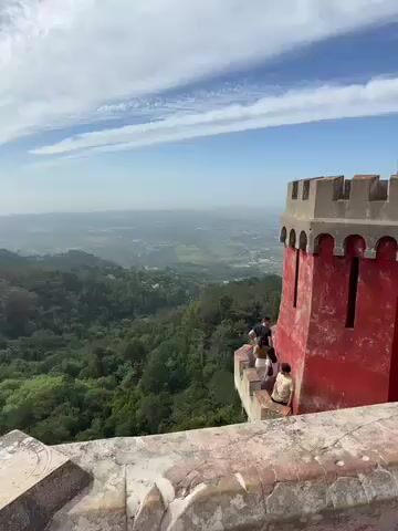 The video captures scenic views from a historic palace perched high above a lush landscape. It opens with a wide shot looking out over a vast, forested valley, with distant towns visible on the horizon under a bright blue sky with scattered white clouds. To the right, a distinctive red castle turret with arched windows and battlements stands out, with several people gathered on its balcony, observing the expansive view. A decorative stone railing forms the foreground.

The camera then pans left, continuing to showcase the dense, green forested hills that stretch as far as the eye can see. The panoramic sweep reveals more of the castle's vibrant exterior, transitioning to a long section of yellow and gray wall, also adorned with battlements. More visitors are seen walking along this wall, taking in the breathtaking scenery.

Finally, the camera tilts down and pans right, revealing a bustling internal courtyard of the palace. The architecture is strikingly colorful, featuring buildings painted in bright yellow and gray, a prominent dome, and a round tower. Many people are leisurely walking and congregating in the courtyard, exploring the historic site. The overall impression is one of a sunny day with good weather, ideal for sightseeing at what appears to be the iconic Pena Palace in Sintra, Portugal. No discernible text is visible in the video.