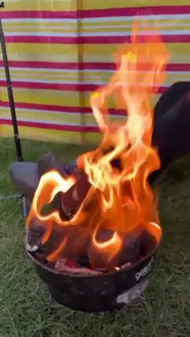 Here is a concise description of the video:

A small black portable fire pit burns with bright orange flames. The fire pit sits on green grass, next to a pair of hiking shoes.  A person wearing a black glove is adjusting the logs in the fire with their hand. Behind the fire pit and shoes is a multi-colored striped wind screen. A metal pole is visible on the left side.