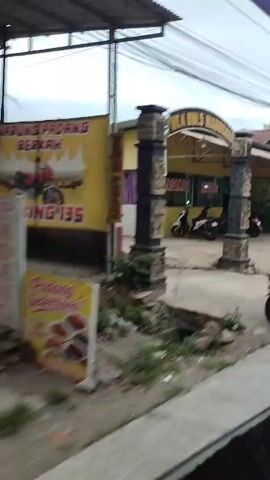 A video shows a person riding on the back of a motorcycle, holding onto the driver. The motorcycle is passing by a building that appears to be a restaurant, with a sign that reads "RM. MINANG". The restaurant has yellow walls and a green trim. There are other motorcycles parked outside the restaurant. The video then pans to show another building with green walls and a wooden door. A person on a motorcycle is seen riding past this building. The video continues to show glimpses of buildings and street scenes.
