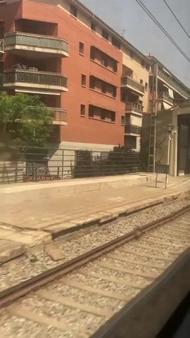 A train is moving past a train station platform. The platform is covered by a roof supported by columns. There are benches and a trash can on the platform. A red brick building is visible behind the platform. The sky is visible above the building and the train tracks are in the foreground. The time stamp indicates the video is about 4 seconds long.