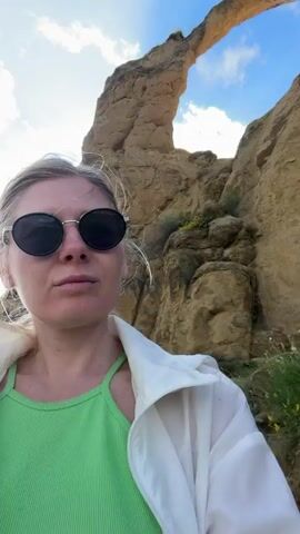 A woman wearing sunglasses and a green tank top stands in front of a rocky, cliff-like formation. She is wearing a white jacket. Behind her, the landscape opens up to a view of a city nestled among green hills under a cloudy blue sky. The video appears to be a selfie-style recording.