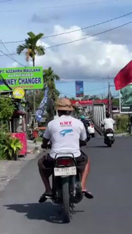 The video shows a man on a motorcycle driving on a road in what appears to be a tropical location. The man is wearing a white t-shirt with the letters "GMT" on it, dark shorts, a straw hat, and sandals. The motorcycle is black and has a license plate that reads "DK 3785 GH". He is following other vehicles along the road. There are signs for a money changer visible on the left side of the road. Palm trees are visible along the roadside, and there are buildings and other structures in the background. The sky is partly cloudy.