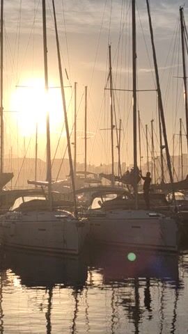 The video shows a harbor at sunset with several sailboats docked. The sun is low on the horizon, shining brightly through the masts and rigging of the boats, casting long shadows on the water. The water is calm, reflecting the boats and the sunlight. A person is standing on one of the boats, silhouetted against the bright sun. The overall scene has a peaceful and serene atmosphere.