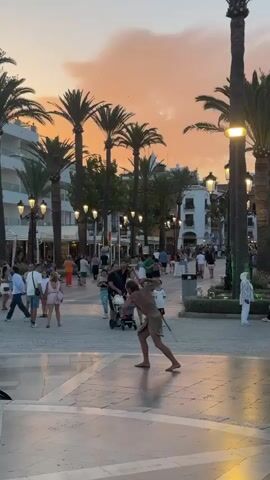 The video captures a lively street scene at dusk, likely in a warm climate, suggested by the prominent palm trees lining the thoroughfare. A street performer, dressed in a light-colored, short-sleeved outfit and shorts, is the central focus, actively engaged in a performance that involves spinning a hoop or similar object around their body. The performer's movements are dynamic and agile.

In the background, a diverse crowd of people strolls along the paved street. They are dressed in casual summer attire, indicating a leisurely atmosphere. Some individuals are walking in groups, while others are alone or with children. The presence of a stroller suggests families are present. Shops and buildings with awnings and lights flank the street, contributing to a vibrant urban environment. The sky above is painted with the warm hues of a sunset, with shades of orange and pink blending with the darkening sky. Streetlights are illuminated, casting a warm glow on the scene. The ground is paved with light-colored tiles, some of which reflect the ambient light. The overall impression is one of a bustling and enjoyable evening in a popular public space.
