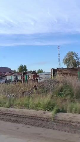 The video shows a roadside view of an outdoor storage area with various construction materials and buildings under a partly cloudy sky. In the foreground, there is a dirt road with tire tracks. In the midground, there is a grassy area with a fence, behind which are stacks of building materials including concrete pipes and what appear to be concrete slabs. Further back, there are several structures, including a long building with a red roof, a smaller wooden house, and a larger industrial-looking building. A telecommunications tower is visible in the distance. A large white billboard with Russian text and phone numbers is also present. The scene appears to be in a rural or semi-rural setting, possibly a construction yard or supply depot. The lighting suggests it is daytime.