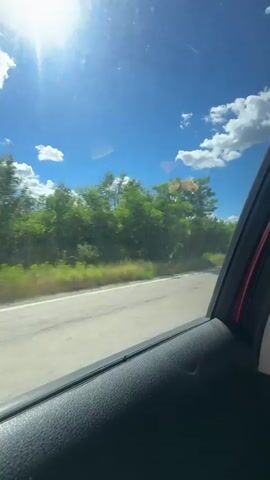 A video shot from inside a vehicle shows a view of the outdoors through the window. The sun is shining brightly in a blue sky with scattered white clouds. Lush green trees and vegetation line the side of a paved road. The interior of the car, specifically the door panel, is visible in the foreground, partially obscuring the view. There are reflections on the car window, including what appears to be a person's arm and hand. The scene suggests a sunny day, possibly a road trip or travel through a natural landscape.