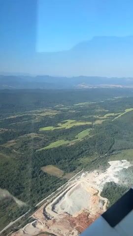 An aerial view from an airplane window shows a vast landscape of rolling green hills and forests under a clear blue sky. In the foreground, a quarry is visible with excavated earth and piles of white material, likely rock or gravel. A road or railway line runs alongside the quarry. The sun casts shadows, suggesting it is daytime. The camera angle is downward and slightly to the right, providing a panoramic perspective of the terrain. The bottom right corner of the image shows a part of the airplane's wing or fuselage.
