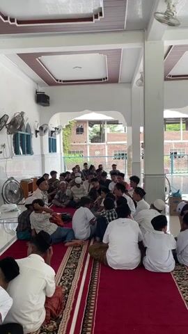 A group of Muslim men and boys are gathered inside a mosque. They are seated on the floor, some on a red carpet with a patterned border. The men and boys are wearing traditional Islamic clothing, including white shirts, some with peacoats, and skullcaps. One man, likely an elder, is speaking to the group. The mosque appears to be an open-air structure with white walls and columns. In the background, there are buildings and trees visible through an open area. Fans are mounted on the walls, and there is a speaker on the ceiling. The lighting appears to be natural and artificial. The overall atmosphere is one of community and religious gathering.
