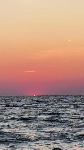 The video captures a sunset over the ocean. The sky transitions from a warm orange hue at the top to a vibrant pink and then a deep magenta near the horizon. The sun is partially visible, a bright pink disc just peeking above the water. The ocean is calm, with gentle waves creating a textured surface. Two faint streaks of clouds are visible in the sky, and a thin white contrail from an airplane cuts across the upper right portion of the frame. The overall scene is serene and picturesque.