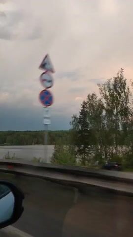 A car drives past a calm lake bordered by a dense green forest on a cloudy day. The sky is a mixture of white, grey, and some patches of blue, with pinkish hues around the edges of some clouds, possibly indicating sunset. The camera is positioned inside the car, showing a portion of the car's side mirror and the guardrail along the road. The car is moving, as evidenced by the blurred motion of the roadside vegetation. Distant buildings are visible along the shoreline. There is music audible in the background.