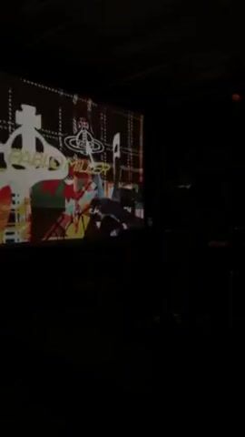 The video shows a dimly lit indoor space where a projector screen is the main source of light. The screen displays a series of animated visuals featuring the name "Pablo Miller" in various graphic styles. In front of the screen, there are shadowy figures of people. The environment appears to be a venue for a presentation or performance.
