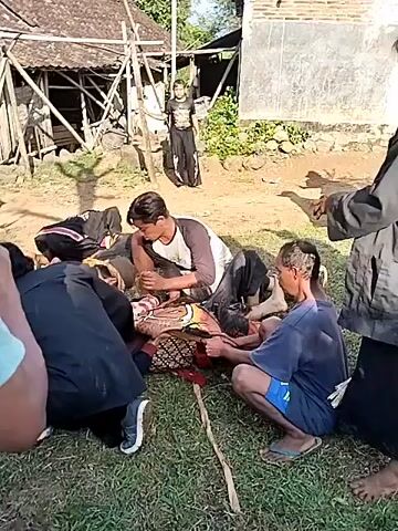 The video shows a group of people gathered in a yard. Most are crouched down or kneeling around a person lying on the ground, covered with a patterned cloth. Several people are seen interacting with the person on the ground, possibly providing assistance or care. The surroundings include a simple building or structure with a wooden frame and corrugated metal roofing, along with another building with a painted exterior. The ground is a mix of dirt and grass. A few individuals are standing and watching the scene unfold. The overall atmosphere seems concerned and focused on the person lying on the ground.