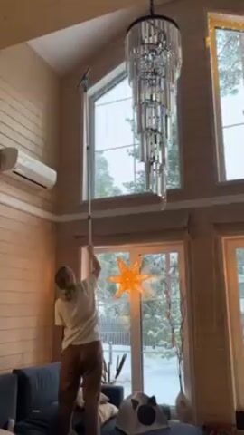Here is a description of the video:
A woman stands on a dark blue sofa in a log cabin with several tall windows and uses a squeegee on an extension pole to clean the tall windows. The chandelier hangs in the middle of the room. There is a lighted star hanging in front of the window behind her. Outside the window, one can see snow-covered ground and evergreen trees.