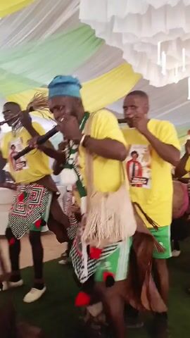 A group of at least nine men in yellow t-shirts and green shorts play instruments and walk forward. The men's shirts have a photo and the number 70 on them. Two of the men are playing long, thin wind instruments, and at least one is playing a drum. The men are standing on green turf in a space decorated with white and yellow fabric. White, modern-looking lights are also suspended from the ceiling.