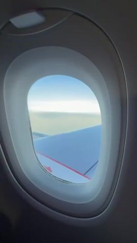 The video shows a view from an airplane window. Initially, the frame includes the window's edge with a view of a white wing with red arrows on it and a horizon line of clouds against a blue sky. The camera then zooms in to focus more on the wing and the sky beyond it. The wing remains the prominent feature, with the clouds and sky filling the background.