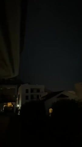 This is a video recording taken at night, capturing a view from a window or balcony. The scene appears to be a residential area with buildings and trees. Several buildings are visible, some with lights on, suggesting they are occupied. There are also parked cars in the foreground. The sky is dark, indicating it is nighttime. There are some clouds in the sky, and one brief, faint flash of lightning is visible in the distance towards the end of the video. The overall mood is dark and somewhat atmospheric, possibly hinting at an approaching storm. The location is identified as Trap, Montenegro. No people are clearly visible, and there is no visible text in the video. The activity appears to be a static observation of the night scene, with the notable event being the flash of lightning.