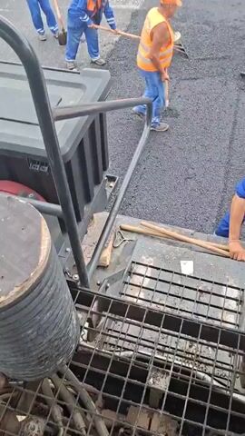 The video shows road construction workers paving a road in Strazhitsa, Bulgaria, on what appears to be a cloudy day. Several men in blue overalls, orange vests, and orange hats are actively involved in the paving process. In the foreground and middle ground, workers use shovels and rakes to manually spread and smooth hot asphalt around a manhole cover and along the road. One worker's orange vest has visible text, possibly "ПЪТНО СТРОИТЕЛСТВО" (Road Construction) in Bulgarian. In the background, two road rollers are seen compacting the newly laid asphalt, moving away from the camera. The setting includes residential buildings, green areas, utility poles, and streetlights lining the road, indicating a town or village environment. Other vehicles, including parked cars, are visible in the distance. The video is likely filmed from the perspective of an asphalt paver or another piece of heavy machinery.