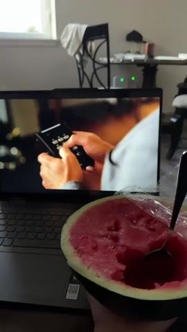 A close-up shot shows a laptop playing a scene from a television show. The screen displays two men, one in a suit and the other in a grey jacket and vest. A half-eaten slice of watermelon with a spoon is in the foreground, obscuring the bottom of the laptop. The background is out of focus but shows a room with furniture and a window. The scene on the laptop shows one man interacting with a device that has buttons. Another scene shows a man in a suit looking to the side. The final scene shows a man in a suit with a stern expression.
