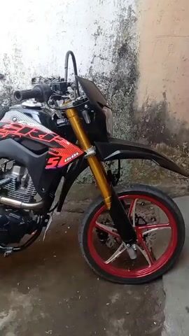 A black motorcycle with red and white graphics is parked in front of a textured wall. The motorcycle has gold-colored front forks, a black fender, and red-rimmed wheels. The seat is black with a textured pattern. The video pans from the front of the motorcycle to the rear, showing the engine, exhaust, and rear wheel. The ground appears to be wet and dirty. There is a piece of clothing hanging on a line behind the motorcycle.