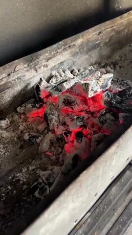 The video shows a close-up, slightly angled view of glowing red embers and charcoal within a metal container. The charcoal is partially covered in white ash. The lighting is dim, with the red glow of the embers providing the primary illumination. The container appears to be a grill or a fire pit. There are no people or other objects visible in the frame. The video is static, with no movement detected.