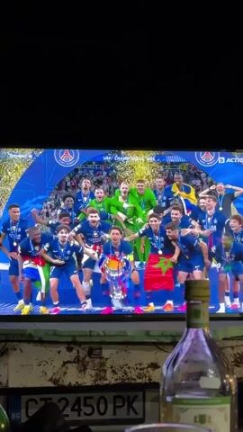 A television screen displays a soccer team celebrating a championship victory, with players in blue and red uniforms hoisting the UEFA Champions League trophy. The words "UEFA CHAMPIONS LEAGUE 2026" are visible above the team. Below the television, a cluttered shelf displays a Bulgarian license plate reading "CT 2450 PK" and a bottle of clear liquid. The overall lighting in the scene is dim, and the television screen is the primary light source. The camera remains static throughout the video.