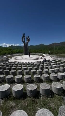 The video shows a wide shot of a monument in a rural outdoor setting. The monument is a large concrete structure resembling abstract arms or stylized flames, standing on a circular platform. Surrounding the platform are rows of many short, cylindrical concrete blocks, likely intended as seating. Two individuals are visible: one standing on the platform near the monument and another sitting on the edge of the platform. The sky is clear and blue.  The background features rolling hills and mountains in the distance.