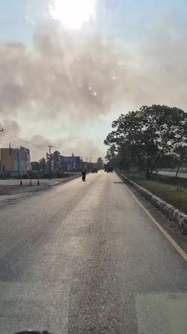 The video shows a view from a vehicle traveling on an asphalt road. The sun is high in the sky, casting a bright glare. Large plumes of smoke are visible in the distance, obscuring the horizon and appearing to be a significant fire or industrial event. On the left side of the road, there are buildings and several orange traffic cones. On the right side of the road, there are trees and a stone wall. Several vehicles, including motorcycles and cars, are visible on the road ahead, suggesting ongoing traffic despite the smoke. The overall atmosphere appears hazy due to the smoke.