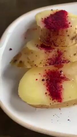 A close-up video shows a white plate with a portion of what appears to be potato slices, topped with a dark red sprinkle, alongside cooked meat in a brown sauce. A small piece of green parsley rests on top of the meat. The meat is cut into pieces and drizzled with a gravy-like brown sauce. The plate sits on a dark brown surface. The video moves slightly, adjusting the focus on the food items on the plate.