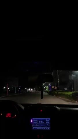 The video shows a car driving down a street at night. Streetlights illuminate the road, and buildings are visible on either side. The camera is positioned inside the car, showing the dashboard, steering wheel, and windshield. The radio is on and tuned to 87.80 MHz. The car appears to be moving slowly and steadily along the road.