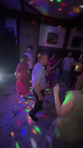 The video captures a lively indoor party scene, likely at night, with a group of people dancing. Colorful disco lights are projected onto the ceiling and floor, creating a festive atmosphere. People of various ages are visible, dressed in casual and semi-formal attire, enthusiastically dancing and interacting.

The setting appears to be a room with dark wooden accents on the ceiling and walls, and framed pictures hanging on the walls. The floor is tiled with a mosaic pattern. The lighting suggests it's a dimly lit environment enhanced by the disco ball or lights. The overall mood is joyful and energetic, with people clearly enjoying themselves. While the exact location is not specified within the video itself, the context provided places it in Zavod Mikhailovskii, Russia. No text is visible in the video.